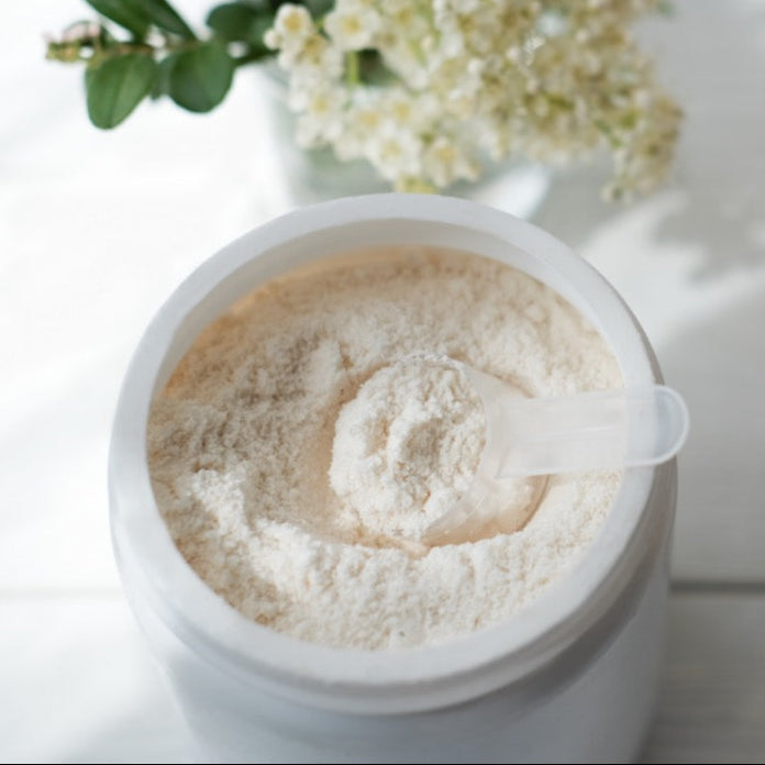 Container of protein powder with a glass of a protein shake on a light surface with flowers in the background