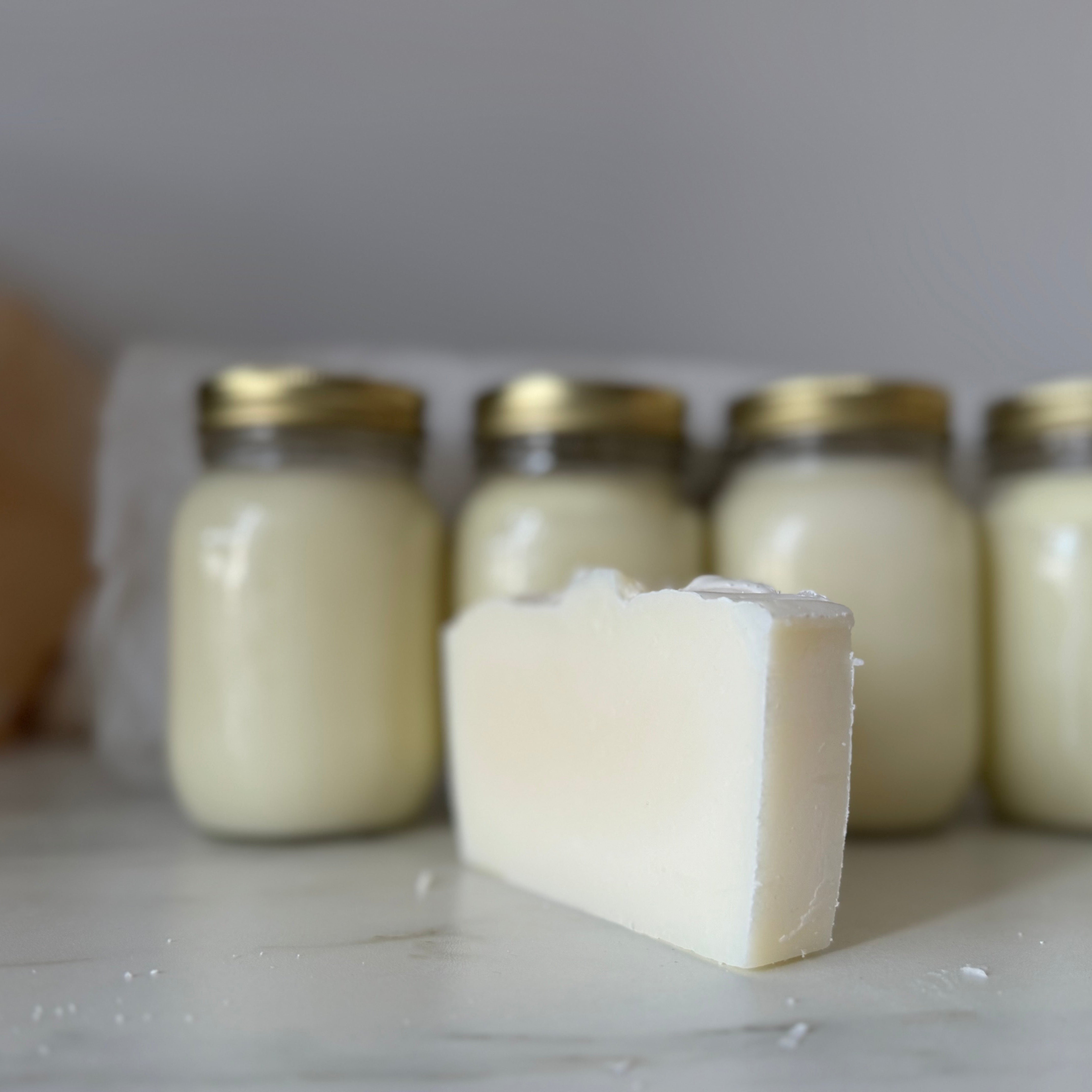 Bar of soap in front of four jars with gold lids on a light surface.
